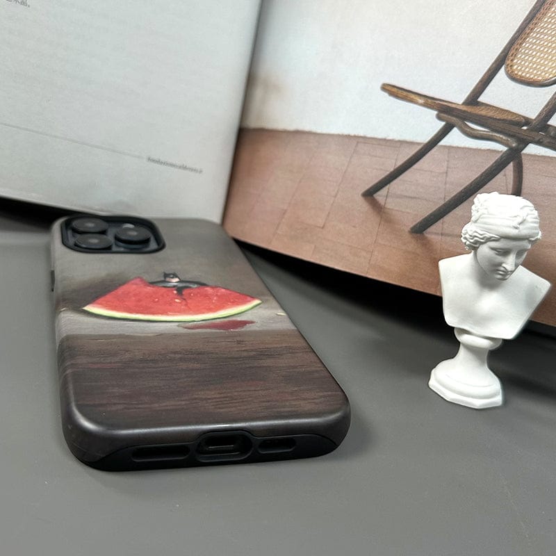 A Quirky Watermelon & Miniature Hero iPhone 16 Pro Max Case featuring a whimsical watermelon slice design, ultra-slim yet offering full protection, is placed on a table. Behind the phone are an open book with a photo of a chair and a small white bust sculpture. The scene is arranged on a gray surface.