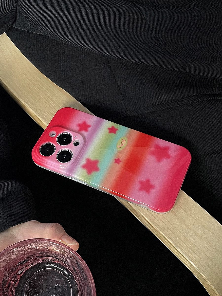 A smartphone with a Pink Starburst iPhone 16 Pro Max Case featuring a pastel rainbow design is placed on a wooden surface. A hand holding a glass of water is partially visible in the bottom left corner.