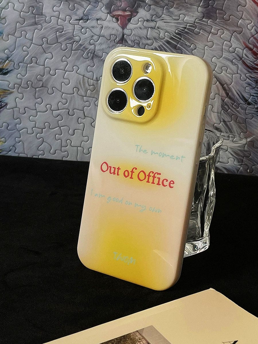 The "Out of Office" iPhone 16 Pro Max case in a gradient yellow design sits upright against a clear support. The stylish all-inclusive protective cover features text at the top with additional writing underneath, while a puzzle and a book are partially visible in the background.