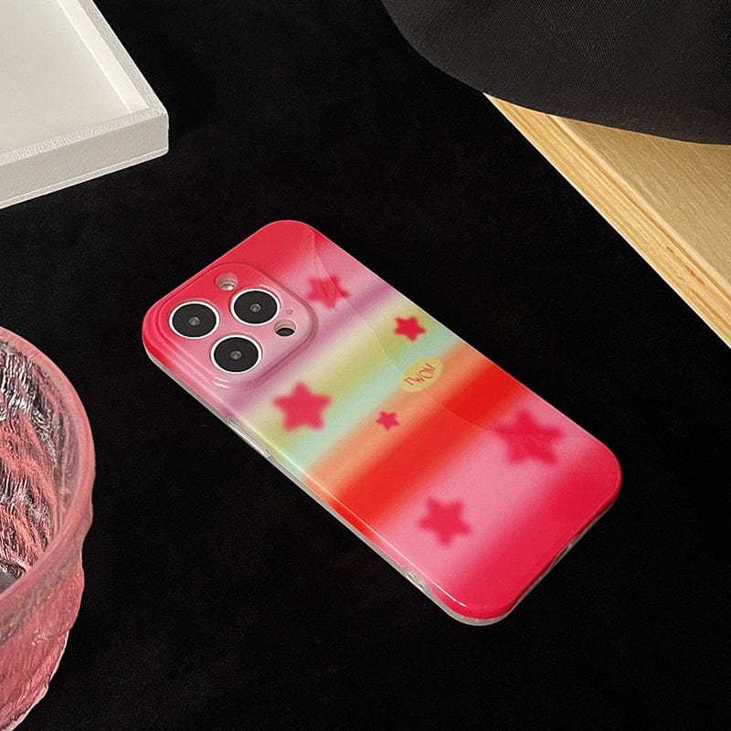 A smartphone lies on a black surface, encased in a Pink Starburst iPhone 16 Pro Max Case featuring a pastel rainbow design with star patterns, near a textured pink item and a beige object.