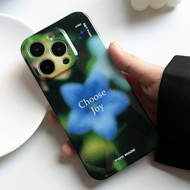 A hand holding an iPhone 16 Pro Max with a "Choose Joy" iPhone 16 Pro Max Case. The case features a blurred floral design, highlighting a prominent blue flower in the center with the phrase "Choose Joy" printed on it. The camera lenses are visible at the top left of the phone.