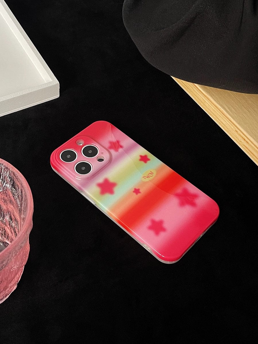 A smartphone adorned with a Pink Starburst iPhone 16 Pro Max Case, which features a pastel rainbow design with red and white stars, is placed on a black surface next to a pink glass bowl and a beige object.