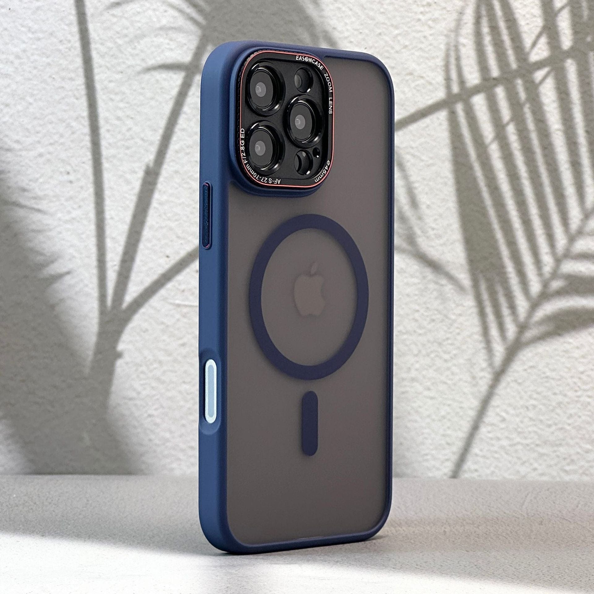 An iPhone 16 Pro Max case with a matte finish and shockproof full-coverage, featuring a MagSafe ring, sits upright against a light-colored surface. The background shows shadows of plant leaves on a light wall, highlighting the sleek design of the dark blue edge protective case.