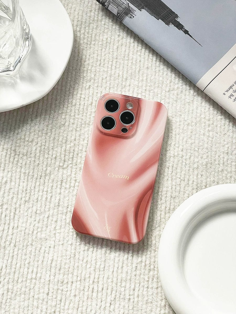 A smartphone with a pink and white marbled Silky Rose iPhone 16 Pro Max case, featuring an elegant marble design and luxurious all-inclusive protective cover, is placed on a textured light-colored surface. A partial view of a glass, a white plate, and a folded newspaper are visible around the phone.