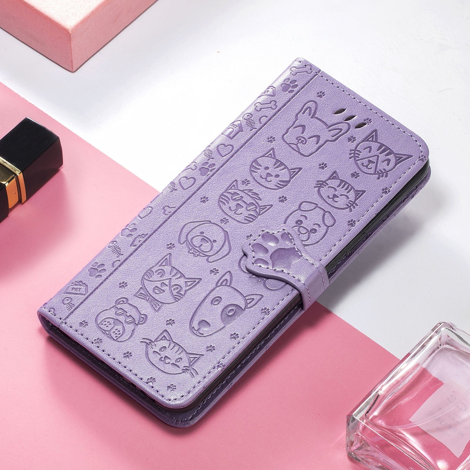 A purple iPhone 16 Pro Max Wallet Case with a PU leather finish and cute cat and dog embossed designs rests on a pink and white surface, accompanied by a lipstick and a clear glass bottle. Perfect for those seeking style and practicality, this wallet case features card slots and a stand.