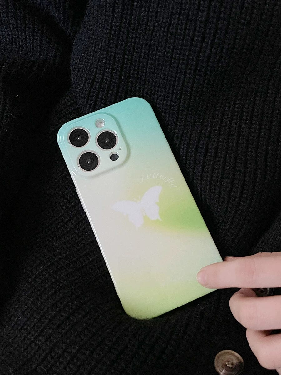 A hand is holding the Butterfly Bliss Gradient iPhone 16 Pro Max Case, which features a soothing green design and a white butterfly motif. The person is dressed in dark-colored, textured fabric.