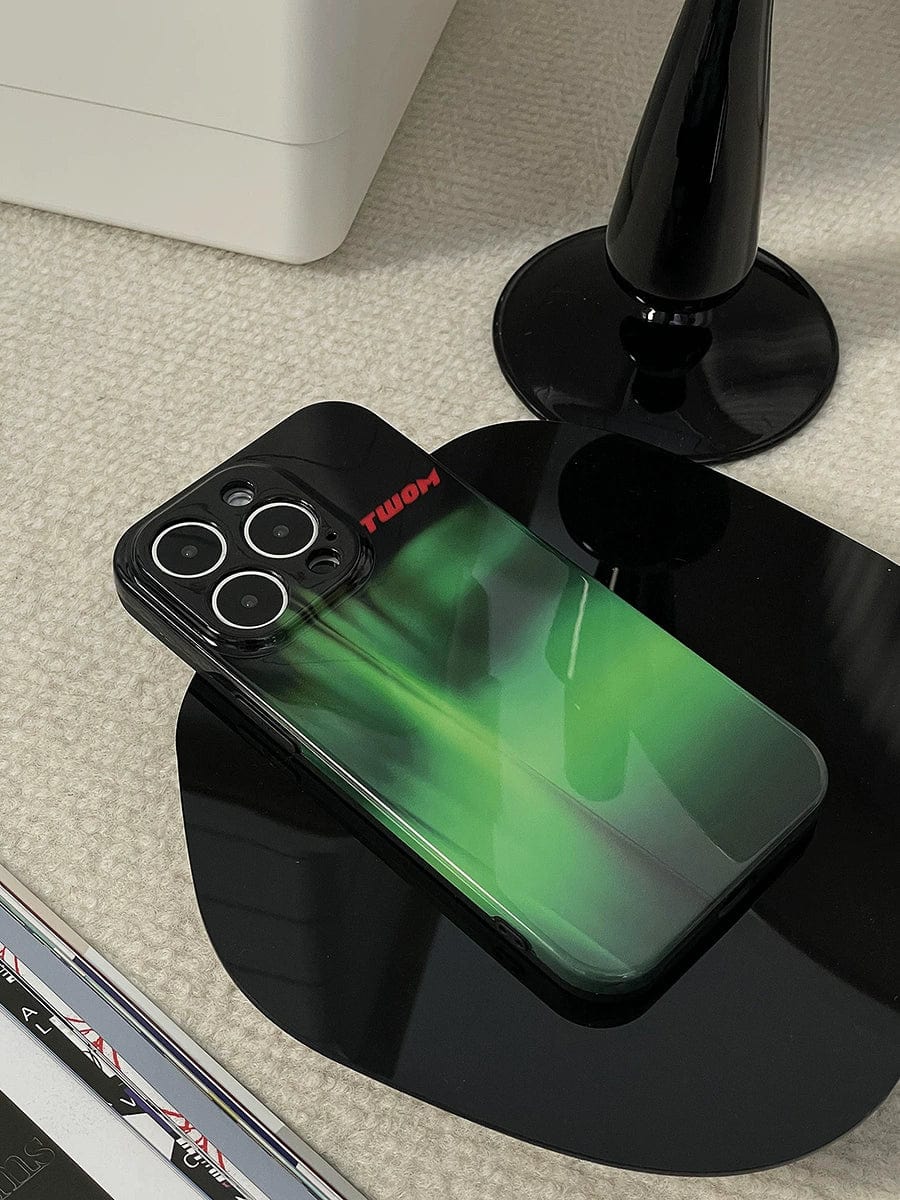 An iPhone 16 Pro Max with an Aurora Green Gradient case featuring a sleek Northern Lights design rests on a black wireless charging pad next to a black lamp base.