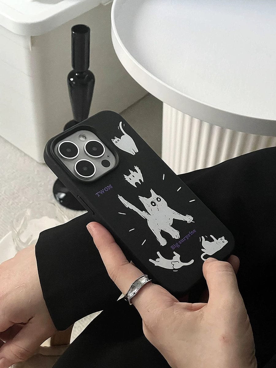 A hand holding a Big Surprise Black Cat iPhone 16 Pro Max case, showcasing playful cartoon cat illustrations in various poses. The background includes a white, round table with a black vase on it, and part of another white object. The person is wearing a silver ring.