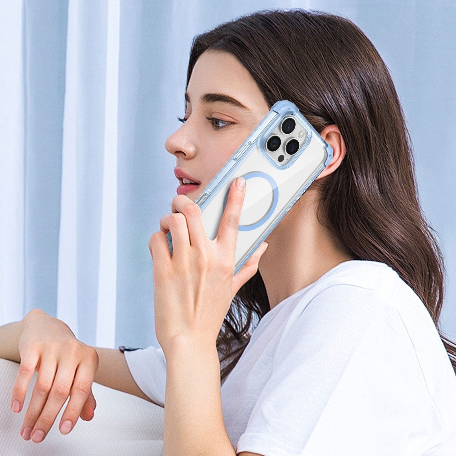 A woman with long dark hair holds an iPhone 16 Pro Max Clear MagSafe Case to her ear while sitting on a white couch, facing left. She wears a white t-shirt and has a neutral expression. The case boasts military-grade drop protection, 3H anti-scratch technology, and is available in multiple colors.