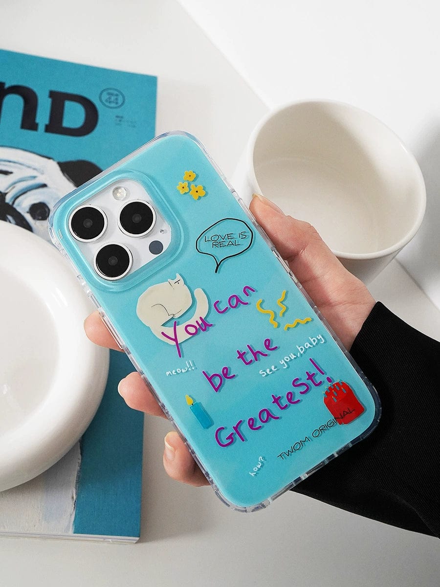 A hand holding the "You Can Be the Greatest iPhone 16 Pro Max Case," which boasts a fun motivational design in blue with colorful doodles and affirmations such as "You can be the Greatest" and "Love is real." In the background, you see a magazine and a white bowl.