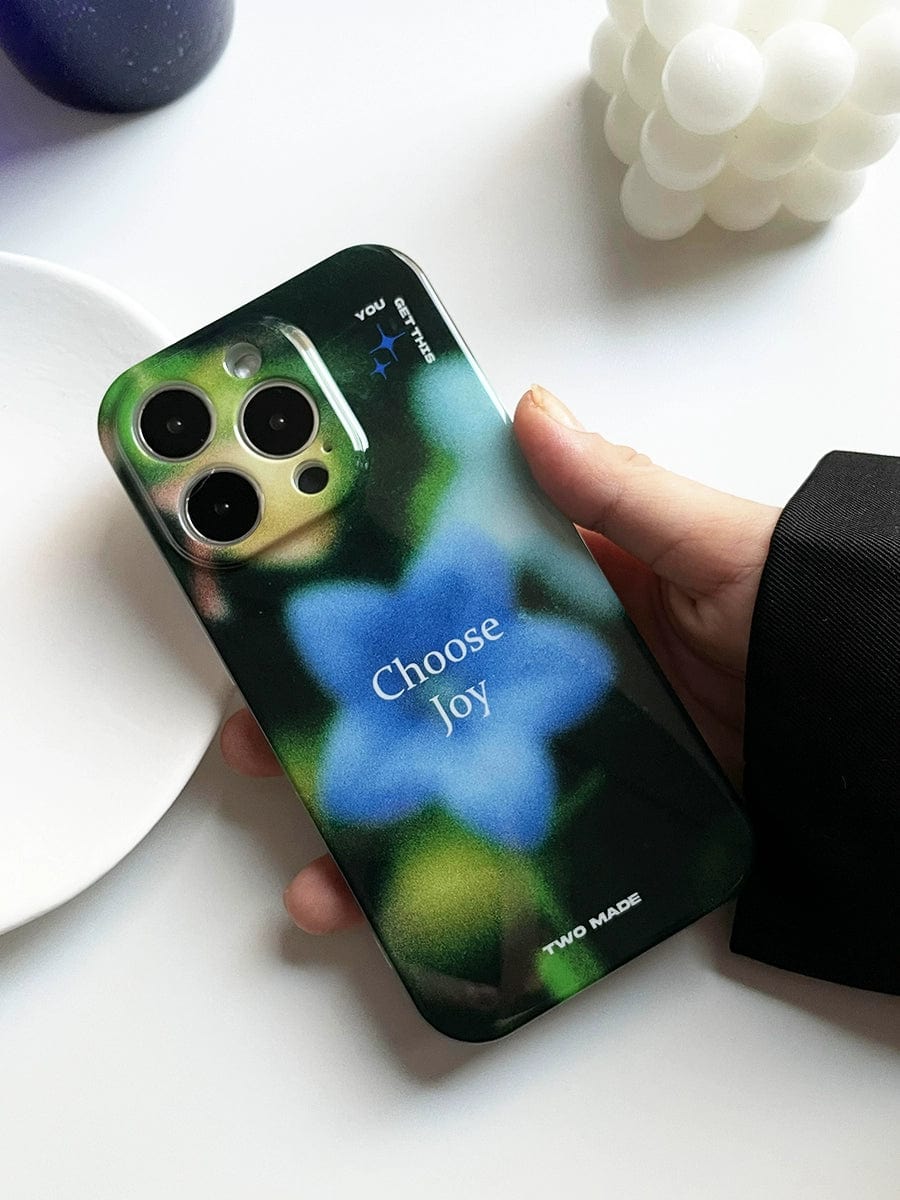 A hand holding the "Choose Joy iPhone 16 Pro Max Case | Floral Blur Design | Inspirational All-Inclusive Protective Cover," which has a dark green design featuring a blue flower and the text "Choose Joy." The phone case also displays additional text and symbols. In the background, other objects, including a white bubble-like ornament and a purple item, are partially visible.