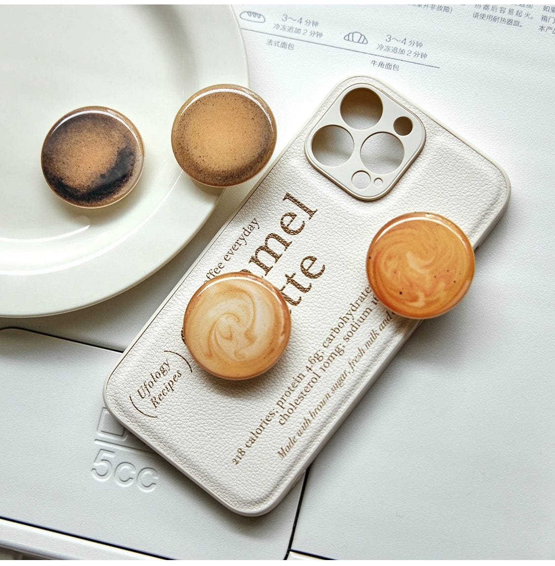 A Caramel Latte MagSafe iPhone 16 Pro Max Case, featuring a coffee-inspired design with "Caramel Latte" text and three coffee-colored, circular grips, is placed on a white surface. Two additional grips that resemble swirls of coffee are resting nearby. A white dish is partially visible in the background.