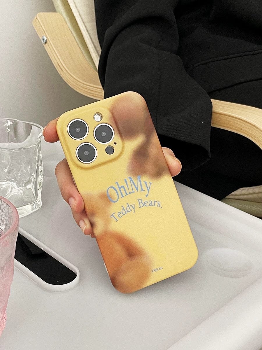 A hand is holding an iPhone 16 Pro Max with the "Oh! My Teddy Bears" iPhone case, featuring a soft matte finish and a delightful yellow and brown design. The phone rests on a light-colored tray next to a pink drinking glass and a clear glass.