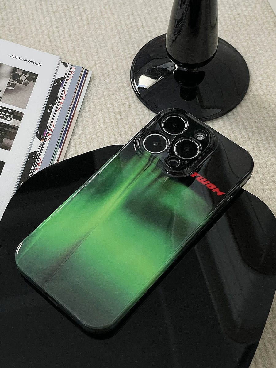 An Aurora Green Gradient iPhone 16 Pro Max Case, featuring a sleek Northern Lights design and an all-inclusive protective cover, lies on a black surface next to a sleek, modern desk lamp and a stack of design magazines.