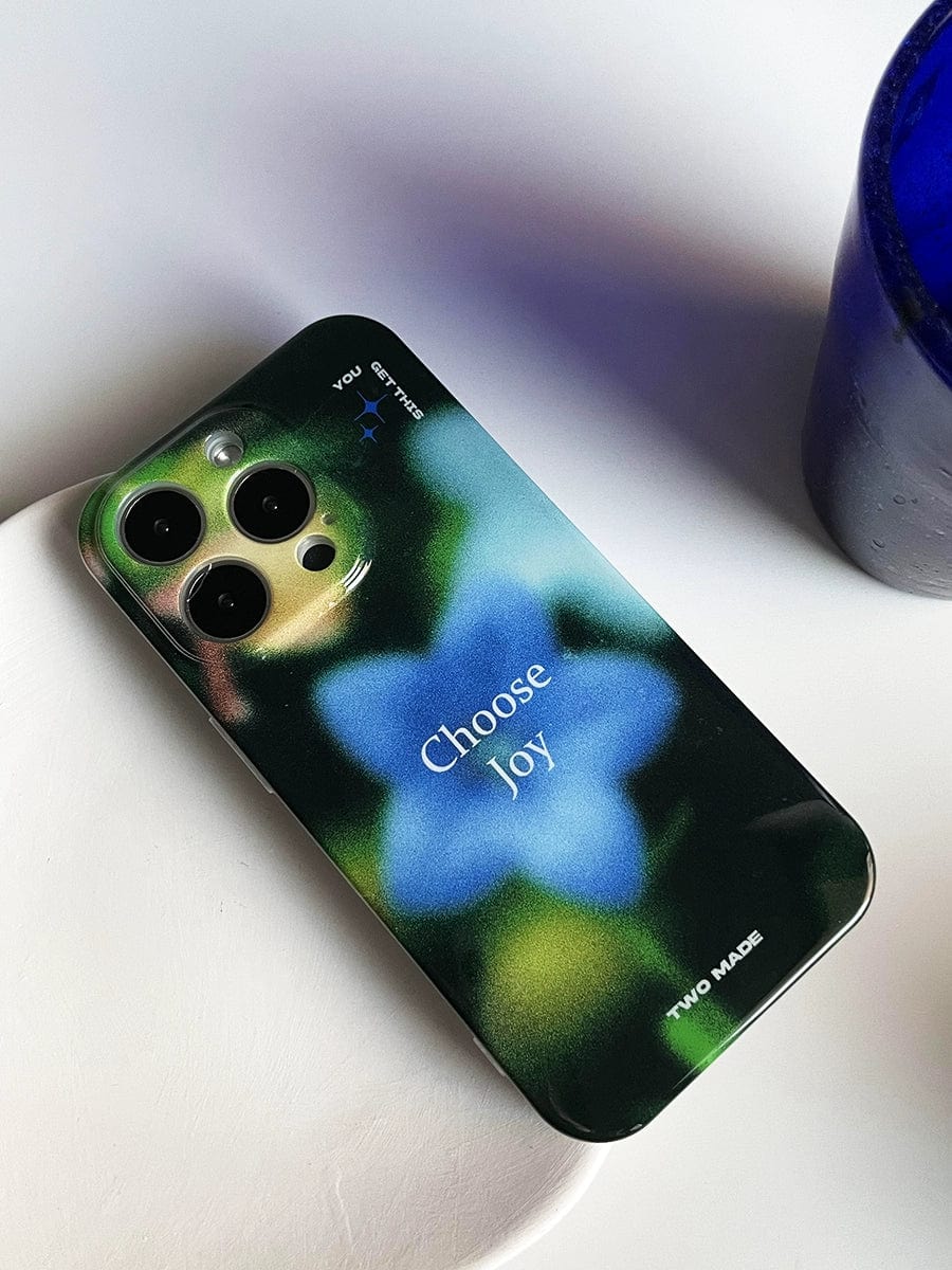 The Choose Joy iPhone 16 Pro Max Case, featuring a blurred green and blue floral design, is placed on a white surface next to a blue container. The inspirational all-inclusive protective cover has the text "Choose Joy" printed on it, along with "YOU" and "ENTER" at the top, and "TWO MADE" at the bottom.