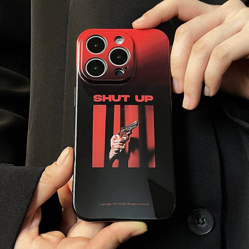 A close-up shows a person's hands holding the "Shut Up Red Gradient iPhone 16 Pro Max Case." The case features a graphic design of a hand holding a gun with "SHUT UP" written above the image. It is primarily black and red, boasting an all-inclusive protective cover with a minimalist, edgy look.