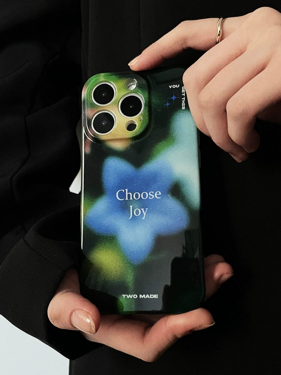 A person in a dark outfit is holding an iPhone 16 Pro Max encased in the "Choose Joy" iPhone case from Two Made, which features a blue-green abstract floral blur design and the printed phrase "Choose Joy." A ring is visible on the person's finger.