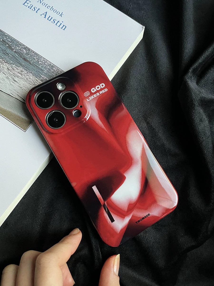 A smartphone with a "God Likes Red" iPhone 16 Pro Max case, featuring a bold abstract design in red and white, is placed on a black cloth. The premium all-inclusive protective cover has text near the camera lenses that reads "LIKE A GOD, LIKE A RED" and there is a book titled "Notebook East Austin" partially visible underneath it.