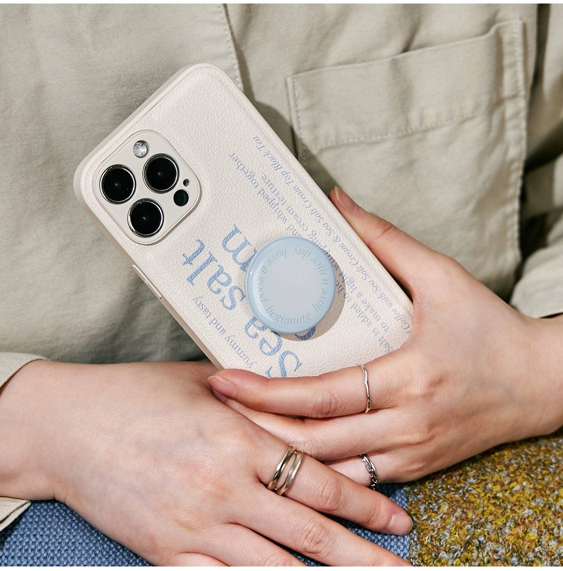 A person holds a smartphone encased in the Sea Salt Cream MagSafe iPhone 16 Pro Max case, boasting a minimalist textured design in beige and featuring a prominent blue circular ring holder. The words "sea salt" are displayed on it. The individual is dressed in a light-colored jacket.