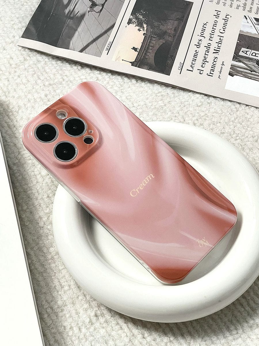 A pink smartphone with three rear cameras is placed on a white, circular charging pad, snug within the Silky Rose iPhone 16 Pro Max Case. The back of the phone has a cream-colored brand name. In the background, there are pieces of a newspaper or magazine on a light fabric surface.