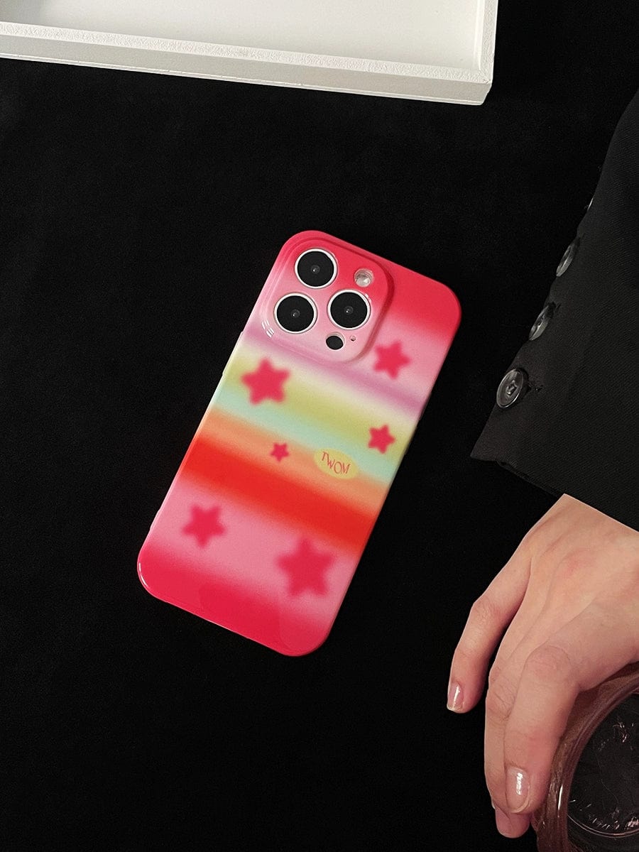 A sleek Pink Starburst iPhone 16 Pro Max Case with a red exterior adorned with pink stars and a pastel rainbow stripe pattern on a black surface. A hand is partially visible next to it.