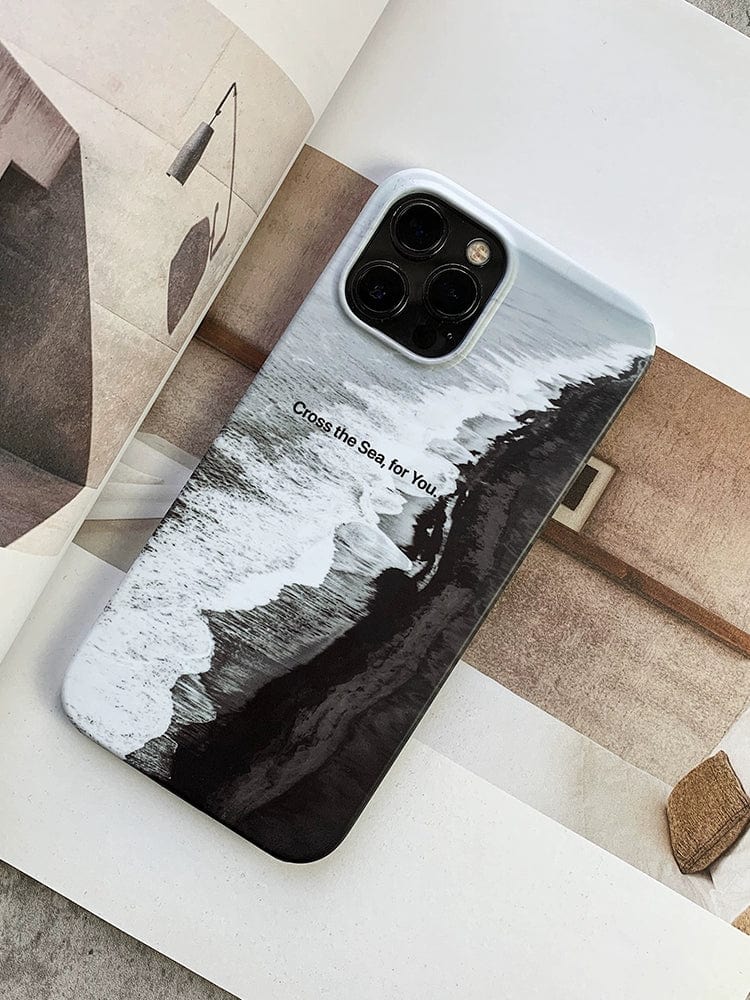 A smartphone adorned with the "Monochrome Ocean iPhone 16 Pro Max Case - Minimalist and Protective," which showcases an aerial view of waves crashing against a shoreline alongside the text "Cross the Sea for You." The phone is placed on an open book displaying abstract, earthy-toned imagery.