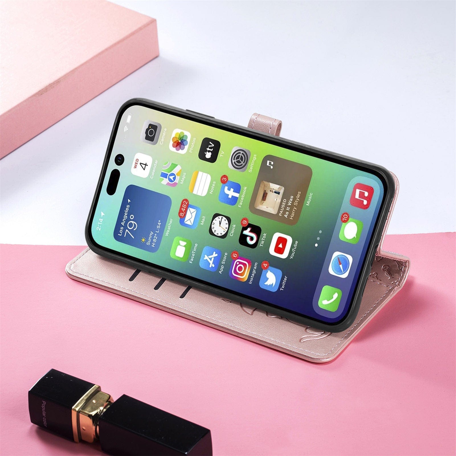 An iPhone 16 Pro Max is propped up in an iPhone 16 Pro Max Wallet Case with a cute cat and dog embossed design, made from PU leather, displaying its home screen with various apps. A tube of lipstick is placed next to it on a pink surface.