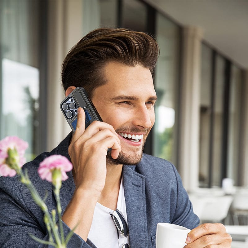 A man sits outdoors, smiling as he holds an iPhone 16 Pro Max equipped with a Full-Coverage Leather Business Case with MagSafe, Rotating Stand, and Card Holder to his ear. In his other hand, he holds a white cup, while pink flowers bloom in the foreground.