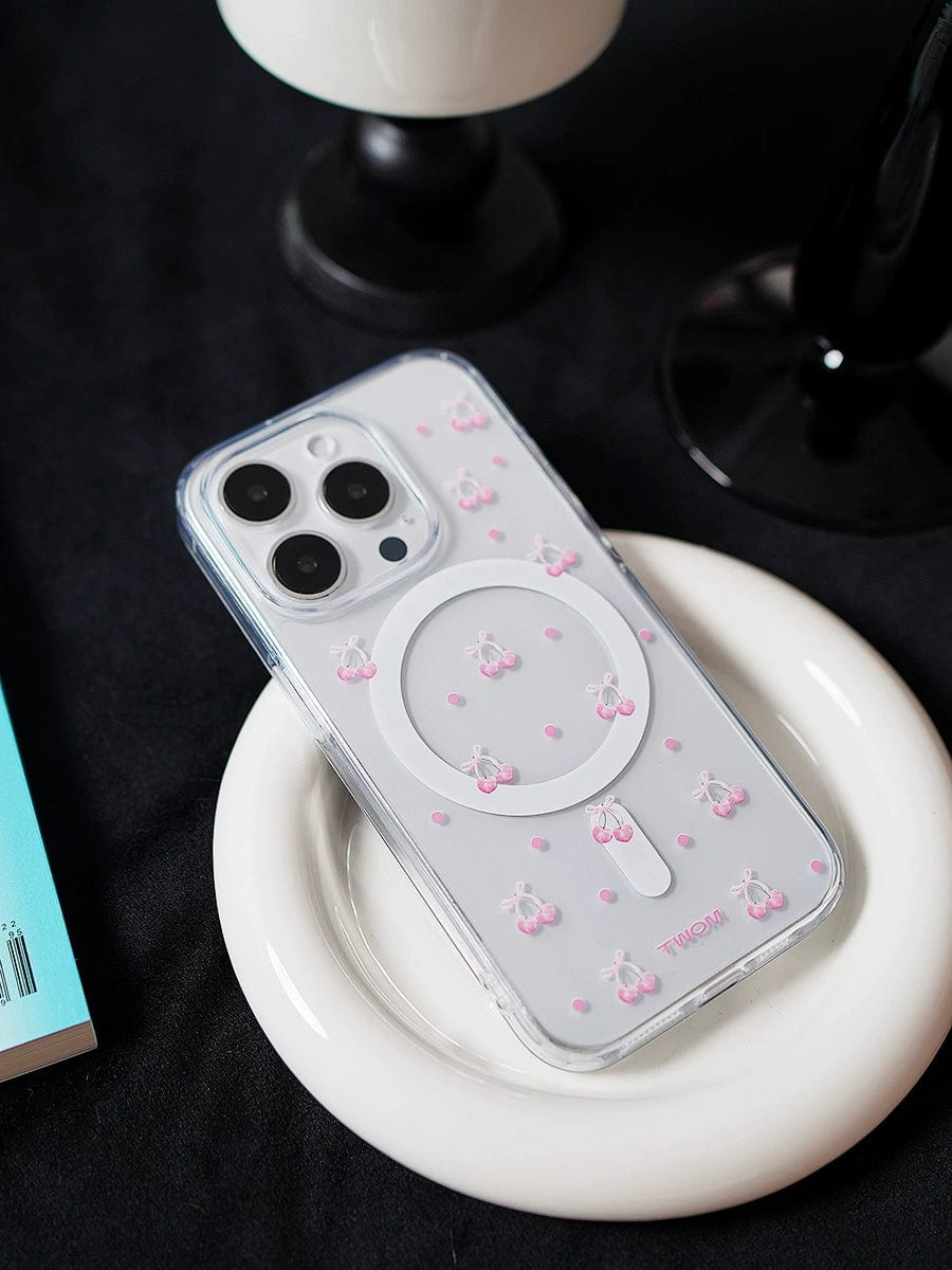 The Cherry Blossom MagSafe iPhone 16 Pro Max Case, featuring a white ring holder and cute 3D pink cherry designs, is placed on a white circular dish on a dark surface. A part of a book with a blue cover is visible to the left, and a black stand or object can be seen in the background.