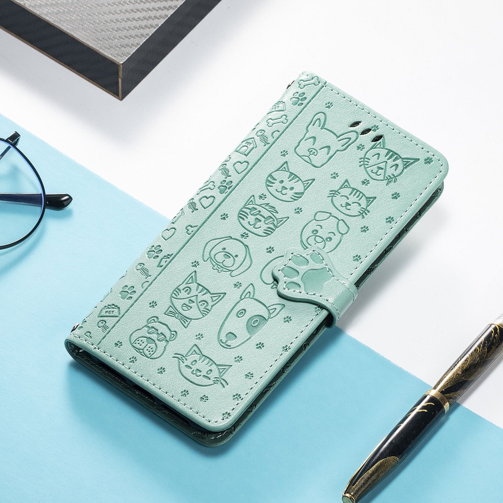 The iPhone 16 Pro Max Wallet Case with Cute Cat and Dog Embossed Design, featuring a light green PU leather flip cover with card slots, stand function, strap, and a paw-shaped clasp, is set on a white and blue surface alongside a pen, glasses, and a notebook.