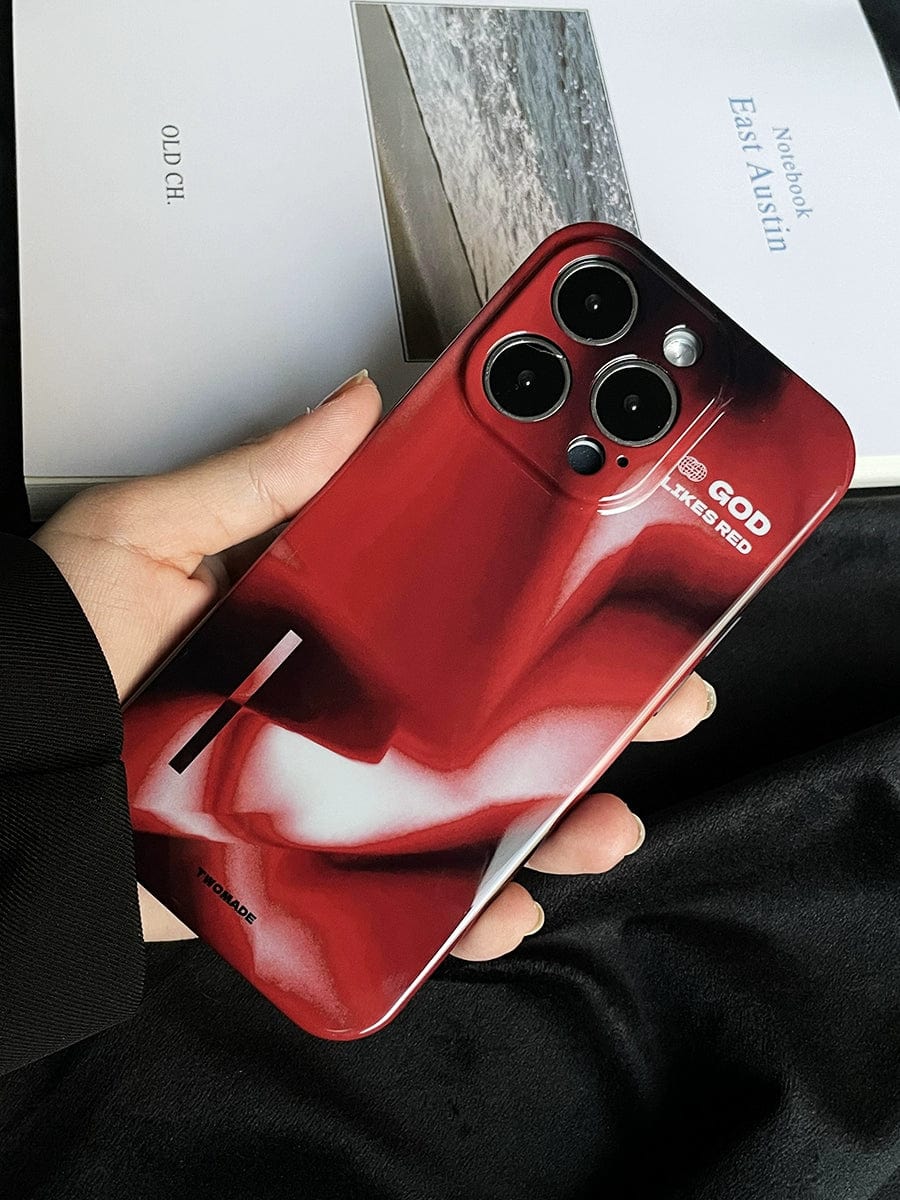 A hand is holding the "God Likes Red iPhone 16 Pro Max Case" with a bold, abstract red and white design on the back. The phone features three camera lenses and has the text "GOD 3/365 RED" inscribed on the premium, all-inclusive protective cover. In the background, there is an open notebook displaying a photo of a beach printed on one of its pages.