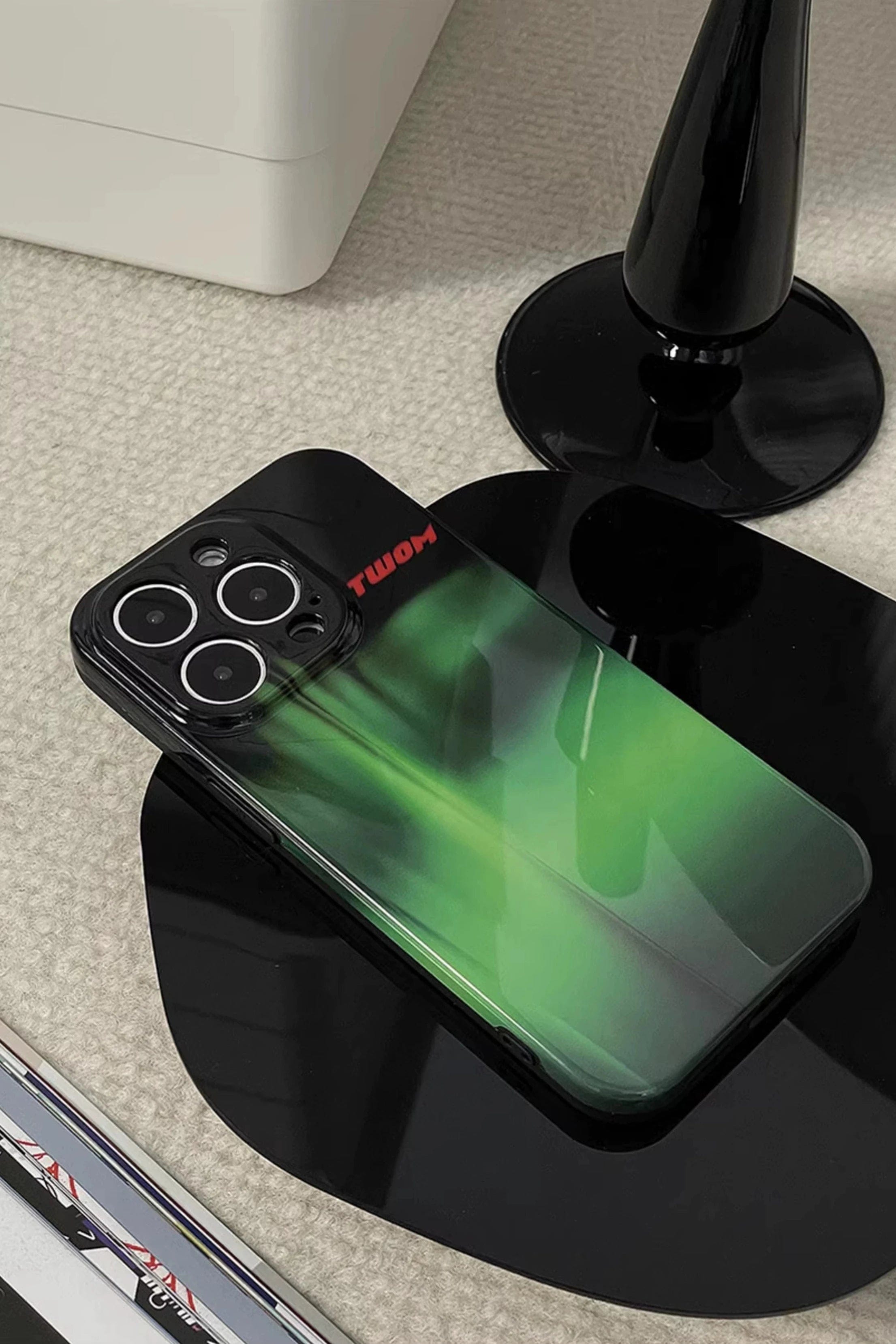 An Aurora Green Gradient iPhone 16 Pro Max Case with a sleek Northern Lights design and the word "TWOONE" is placed on a black surface next to a sleek, black vase.