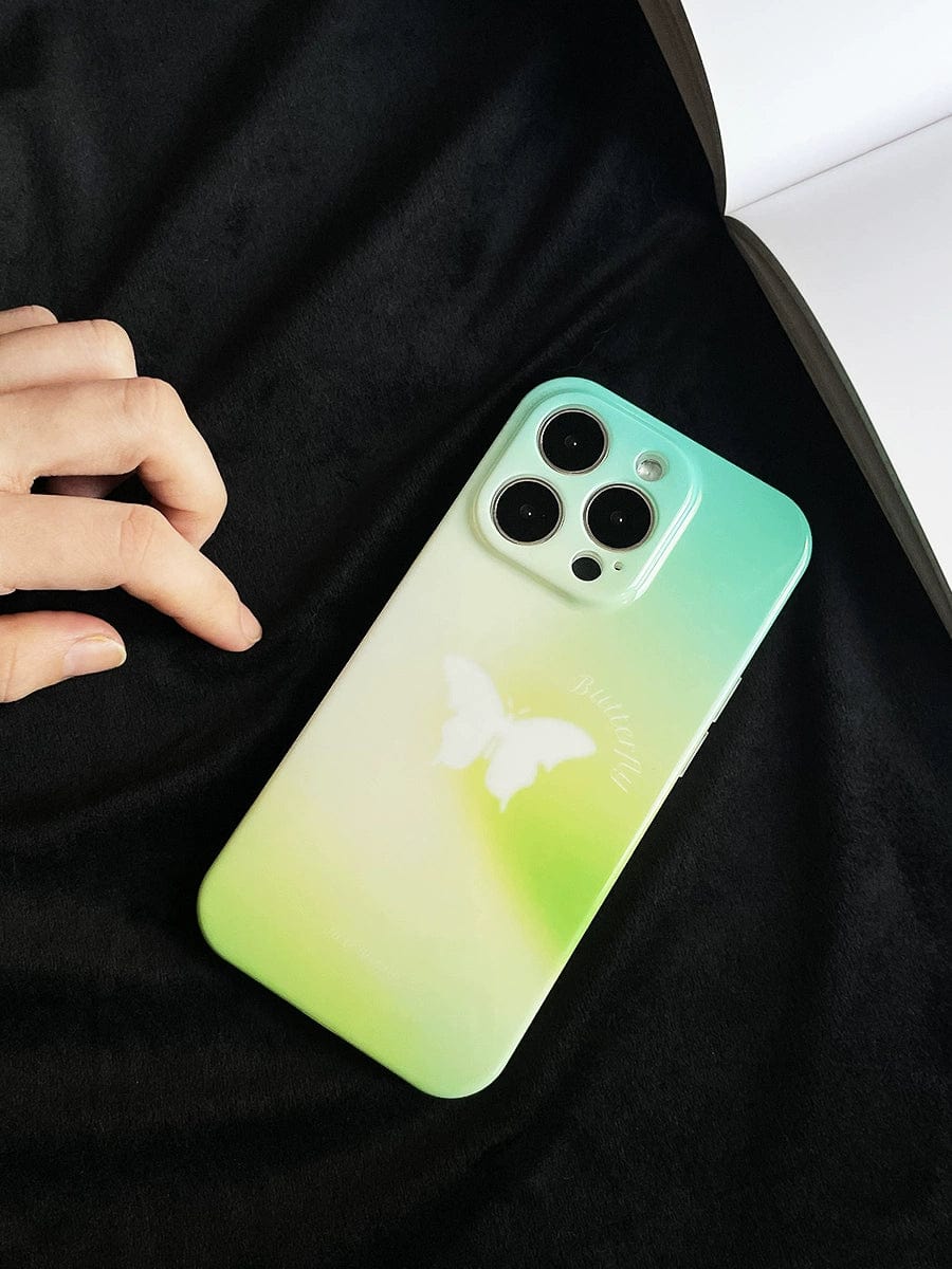 A hand reaches for the Butterfly Bliss Gradient iPhone 16 Pro Max Case, featuring a soothing green design with a white butterfly motif, lying on a black surface next to an open book.