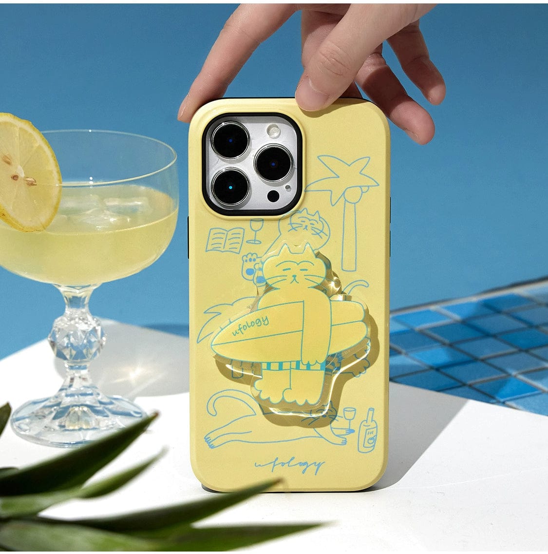 A hand places the Tropical Cat MagSafe iPhone 16 Pro Max Case, featuring a sunny yellow design with a relaxed cat stand by a pool, onto a surface. Next to it is a cocktail glass garnished with a lemon slice. The background includes hints of water tiles and a sky-blue backdrop, suggesting a summer or poolside vibe.