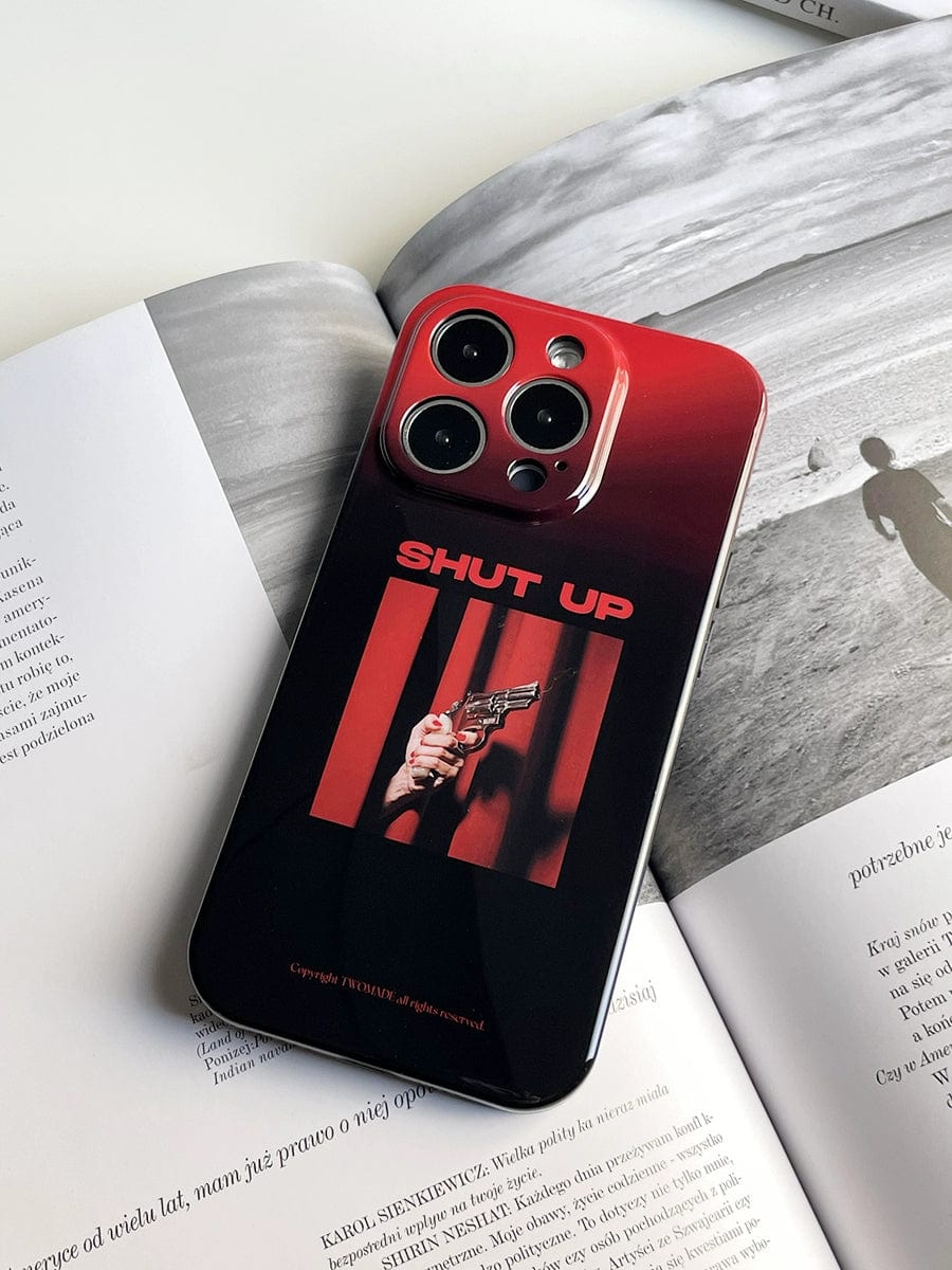 A smartphone with a Shut Up Red Gradient iPhone 16 Pro Max Case lies on an open book. The all-inclusive protective cover features an image of a hand holding a gun and bold red text "SHUT UP" at the top. The phone has a triple camera setup.