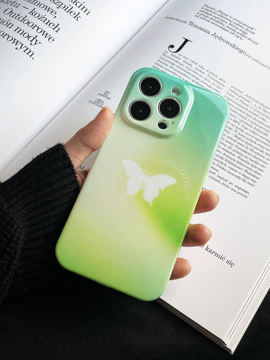 A hand holding an iPhone 16 Pro Max with a Butterfly Bliss Gradient Case in soothing green featuring an elegant butterfly design. The background shows an open book with text.