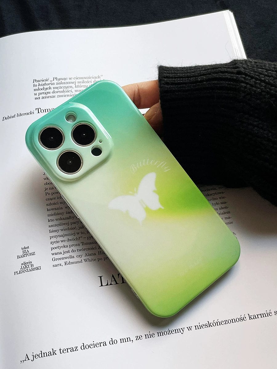 A hand holds the Butterfly Bliss Gradient iPhone 16 Pro Max Case with a soothing green design and white butterfly pattern over an open book.