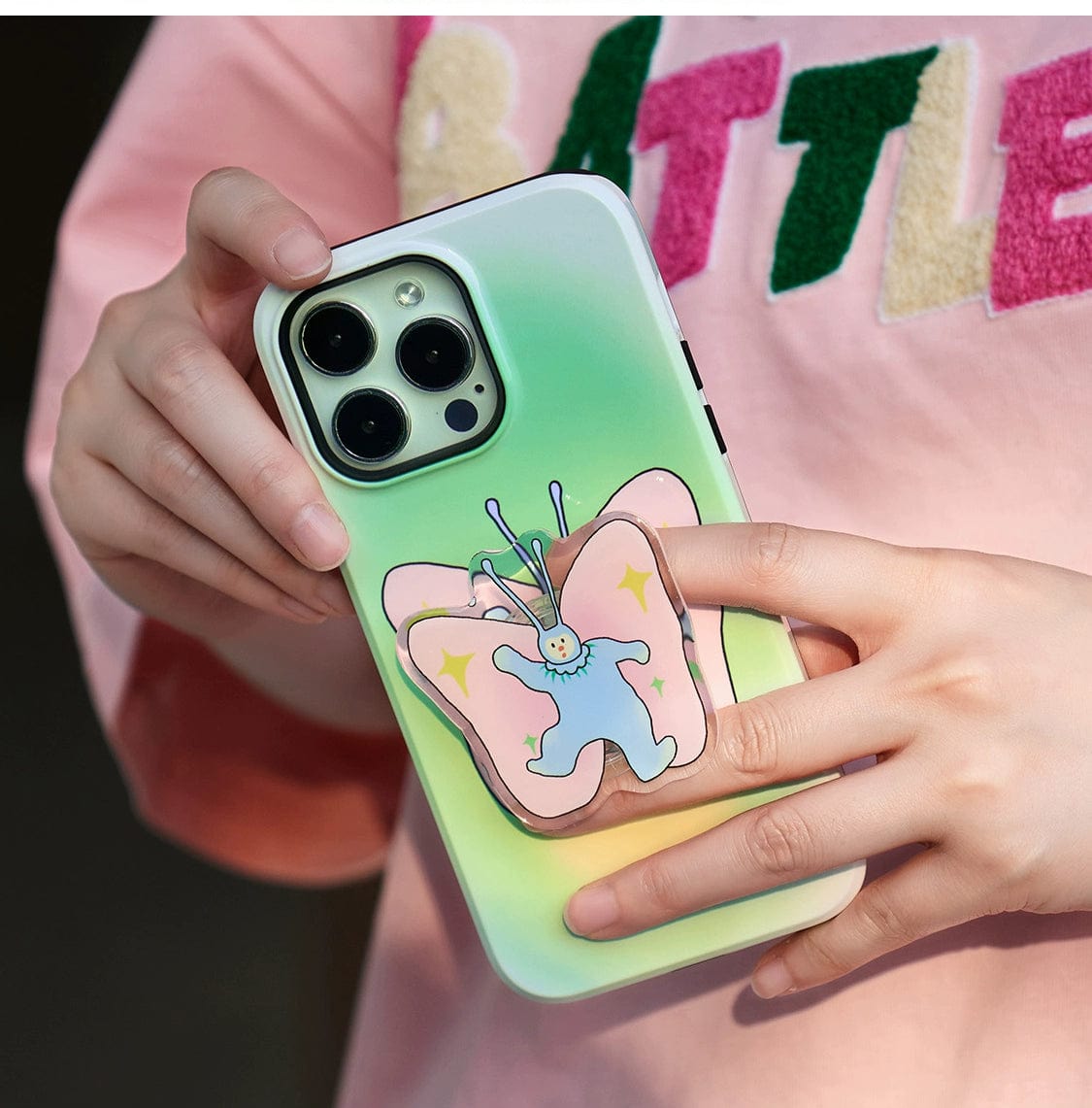 Close-up of a person holding a Golden Sunset MagSafe iPhone 16 Pro Max Case in green, adorned with a decorative butterfly accessory featuring a cartoon character in the center. The person is wearing a pink shirt with partially visible colorful text.