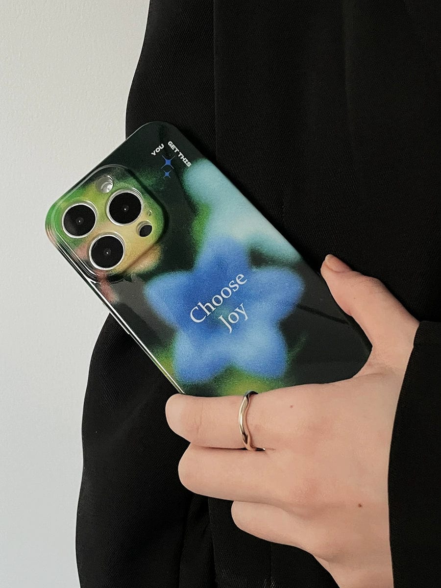 A hand with a silver ring holds the "Choose Joy iPhone 16 Pro Max Case" featuring a floral blur design and the words "Choose Joy." The person is wearing a black garment.