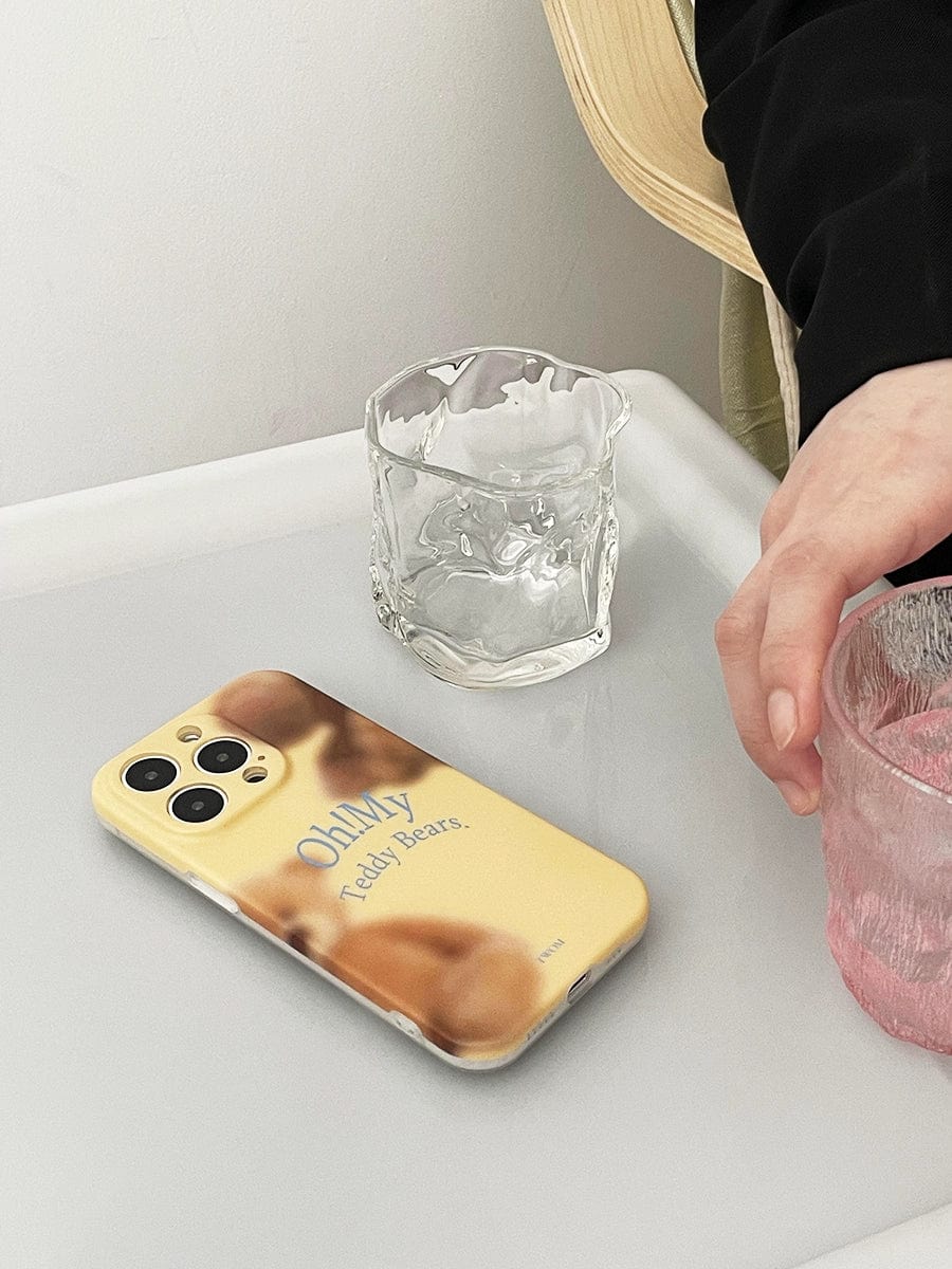 A hand reaches for a pink glass on a white table. Nearby, an Oh! My Teddy Bears iPhone 16 Pro Max Case with a yellow design featuring the text "Only Teddy Bears" and a blurry bear rests on the modern smartphone. An empty, textured glass is placed beside the phone.