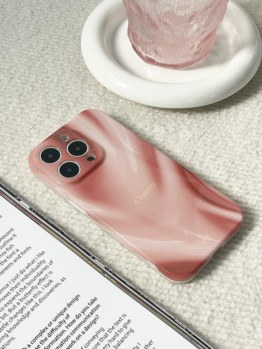 A "Silky Rose" iPhone 16 Pro Max case featuring an elegant marble design, labeled as "Cream," lies screen-down on a soft beige surface beside an open book. In the background, a textured glass rests on a smooth, white circular coaster.