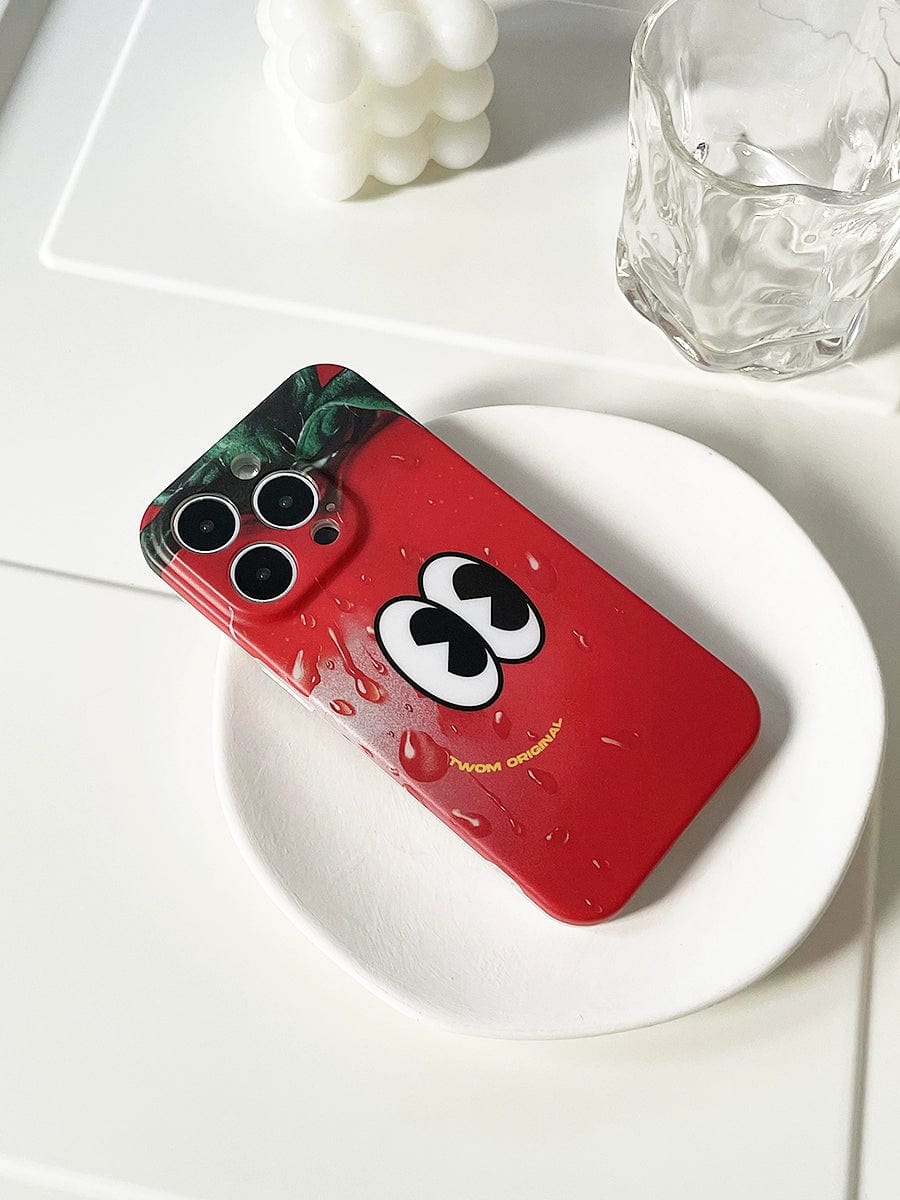 A Tomato Face iPhone 16 Pro Max Case with a fun red cartoon design featuring quirky eyes and water droplets lies on a white plate, next to a glass of water and a decorative object.