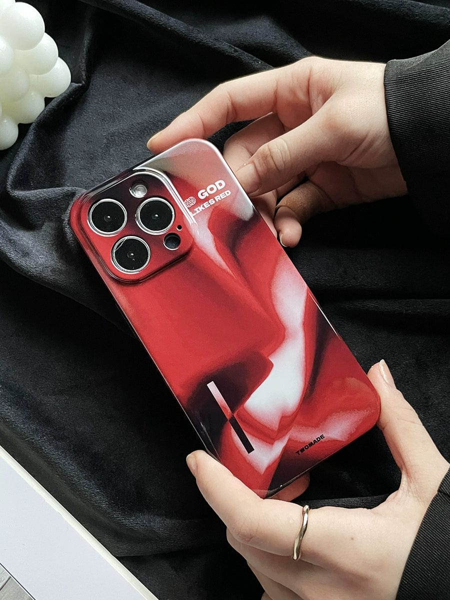 Hands are holding an iPhone 16 Pro Max with the "God Likes Red" premium all-inclusive protective cover, featuring a bold abstract design with the words "God is in his heaven all's right with the world." The phone, equipped with a three-lens camera module, is set against a black fabric background.