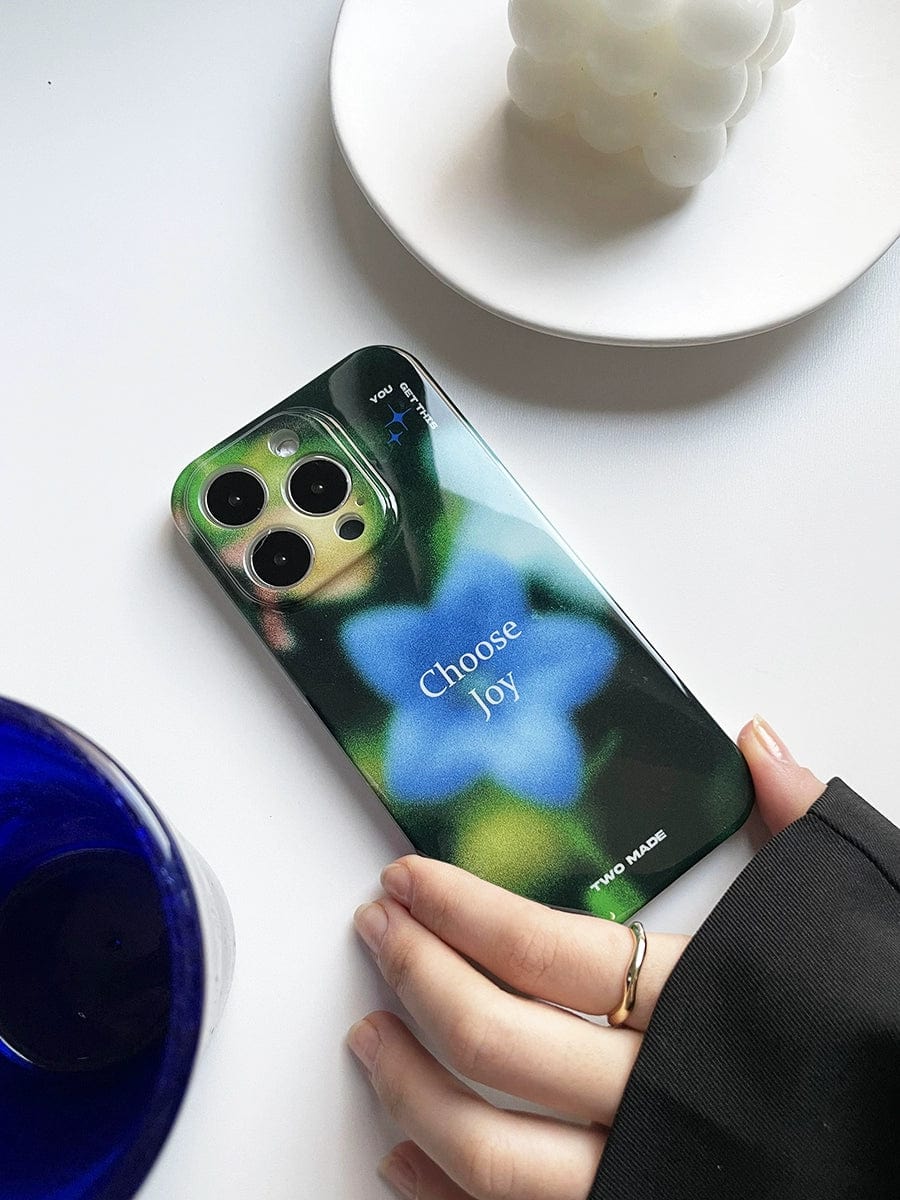 A hand with a ring on the index finger holds an iPhone 16 Pro Max adorned with a "Choose Joy" case from the Inspirational All-Inclusive Protective Cover series, featuring a green and blue floral blur design. The phone rests on a white surface beside a dark blue glass and a textured white candle placed on a white plate.