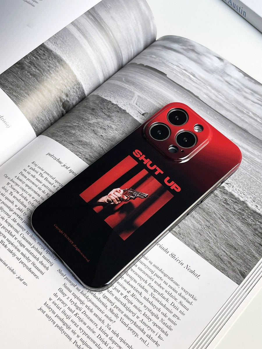 A smartphone with a Shut Up Red Gradient iPhone 16 Pro Max Case, featuring the bold statement "SHUT UP" and an image of a person holding a gun, is placed on an open book with black and white photos and text.