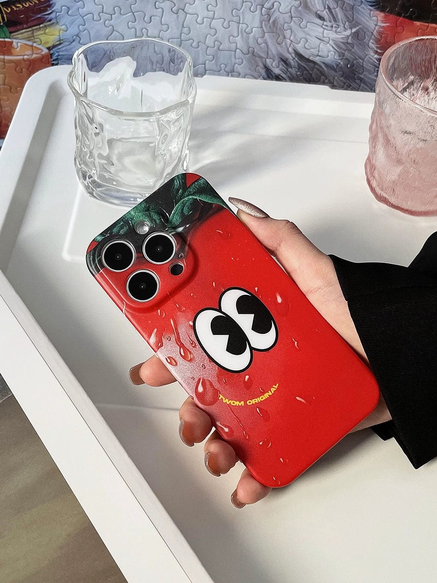 A person holds an iPhone 16 Pro Max with a Tomato Face iPhone 16 Pro Max Case, featuring a fun red cartoon design with eyes and droplets. The phone is next to a glass and a pink container on a white tray.