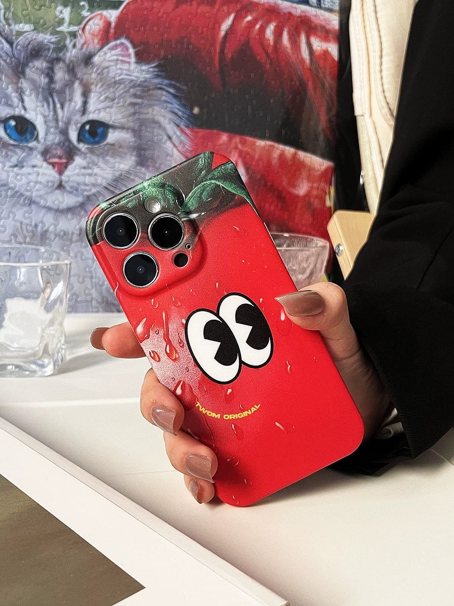 A hand holding an iPhone with the Tomato Face iPhone 16 Pro Max Case, showcasing a whimsical red design with cartoon eyes and a green top, resembling a strawberry. In the background, a painting of a cat adds charm. The new case stands out with its playful and quirky design.