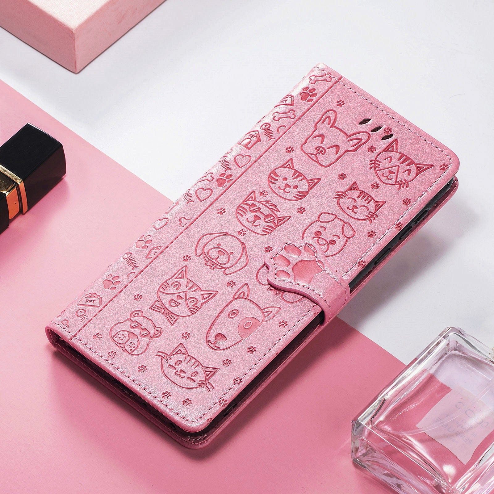 An iPhone 16 Pro Max wallet case, featuring a pink PU leather flip cover adorned with an embossed cat and dog design, lies on a pink and white surface. Nearby are a lipstick case and a clear glass bottle.