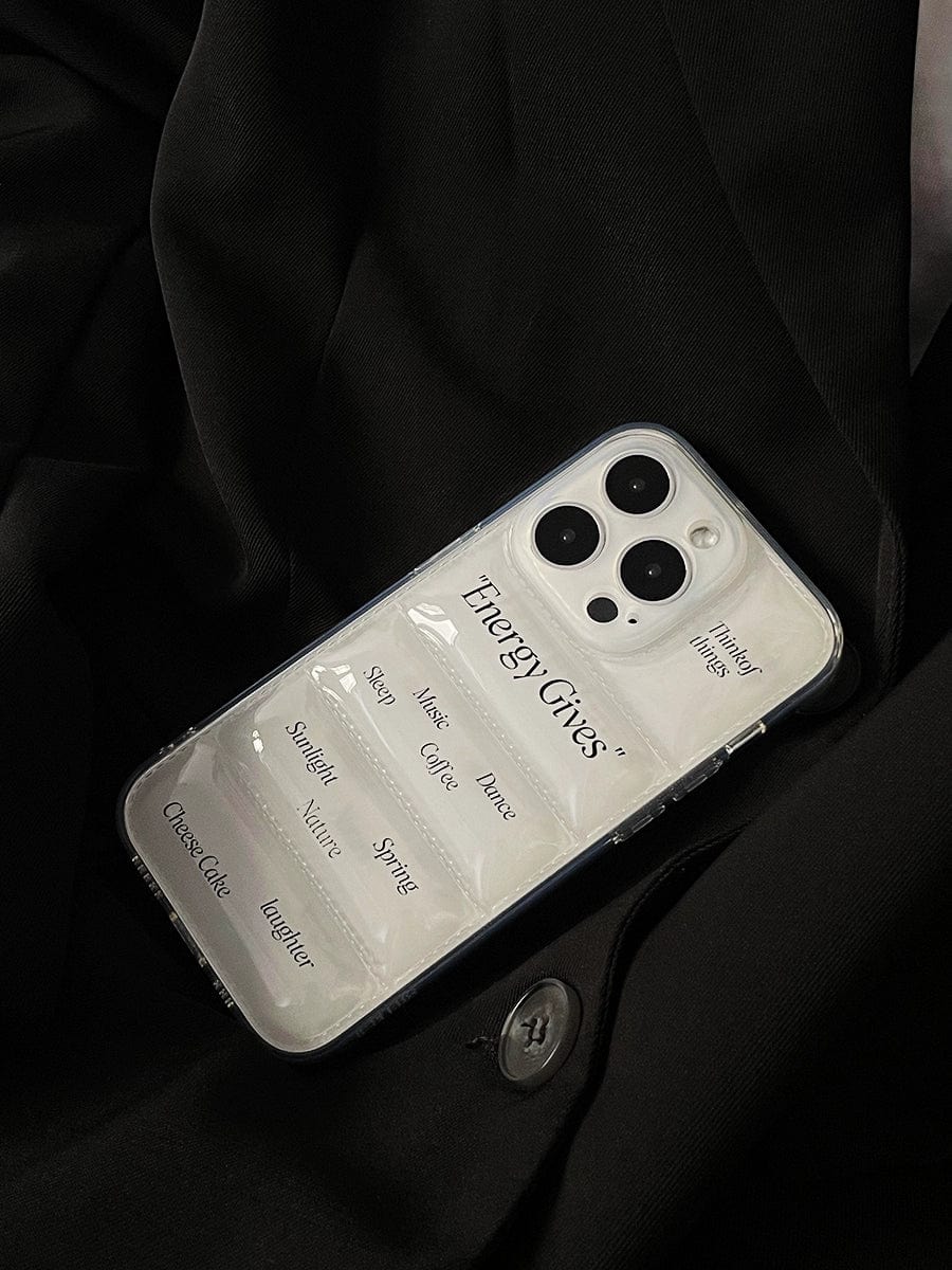 A close-up of the Energy Gives iPhone 16 Pro Max case, featuring a transparent motivational design with the words "EnergyGives," "Mood," "Mental Well," "Sleep," "Nature," "Coffee," "Weather," "Spring," and "Cheese Cake." The phone rests on dark, textured fabric with a visible button.