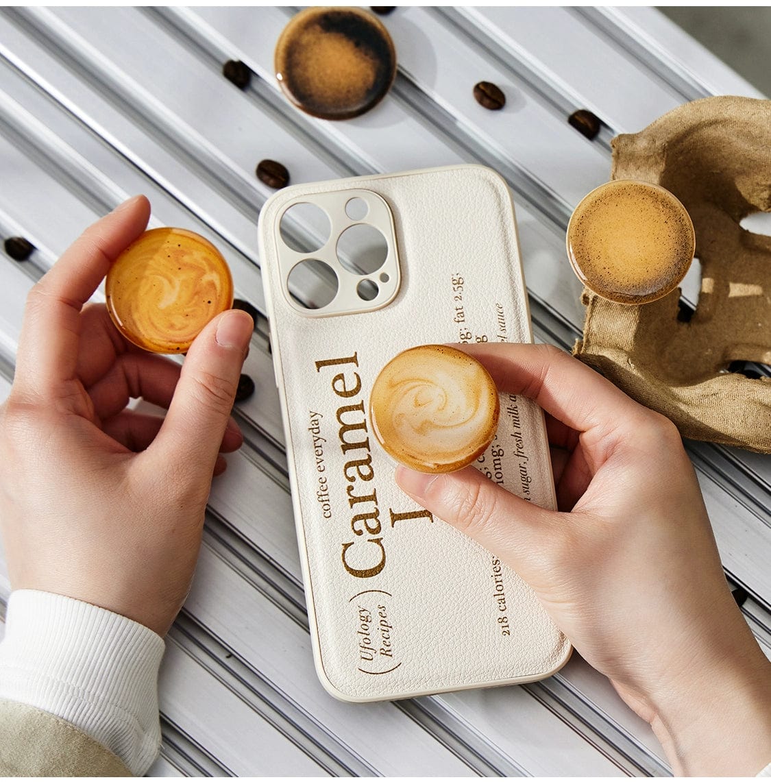 A pair of hands holding a latte art pop socket and an iPhone with a beige Caramel Latte MagSafe iPhone 16 Pro Max Case featuring additional text. Nearby, another latte art pop socket, a cardboard coffee cup tray, and the Caramel Latte MagSafe iPhone 16 Pro Max Case rest on a striped metal surface with coffee beans scattered around.